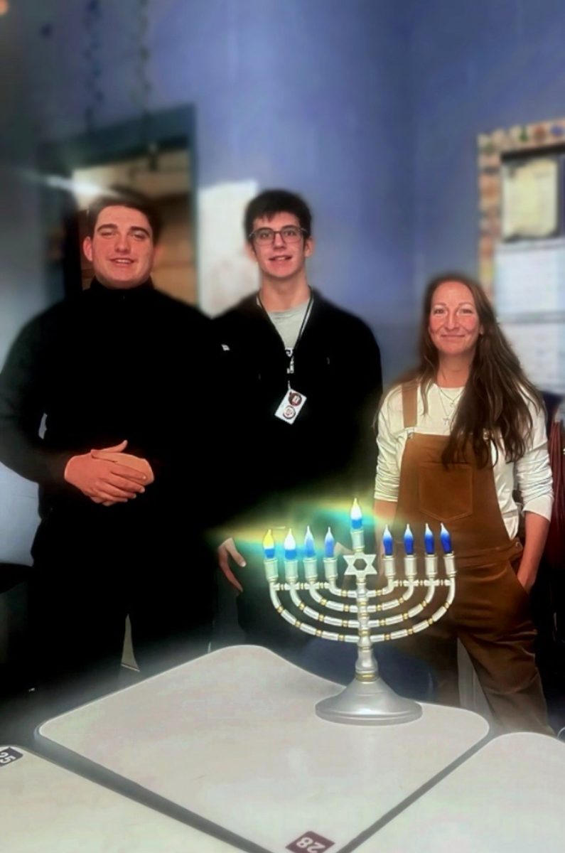 Mark Rozenberg (12), Seth Willgard (11), and Ms Simon stand together in the classroom as they highlight the menorah during the Hanukkah party.